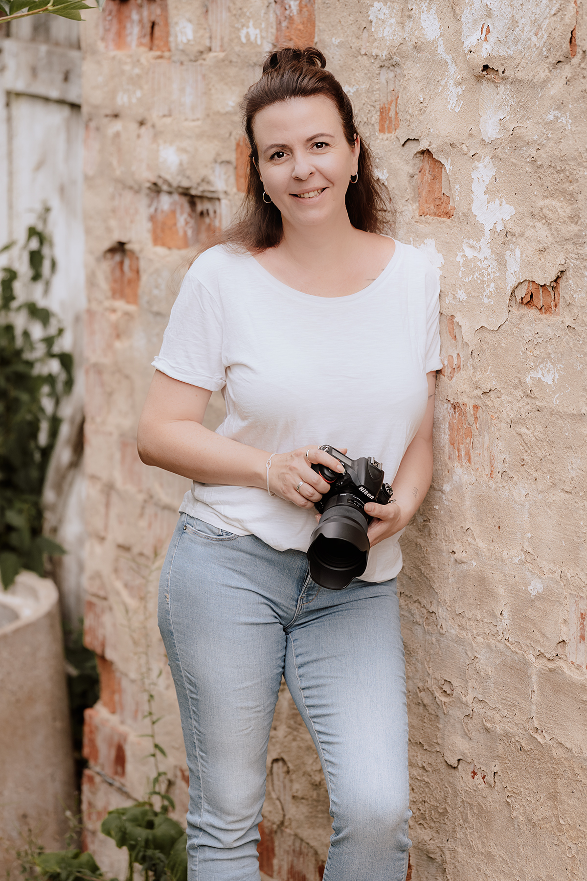 katja-jacobs-fotografin-portraet Katja Jacobs steht lächelnd mit Kamera in der Hand vor einer verwitterten Ziegelmauer