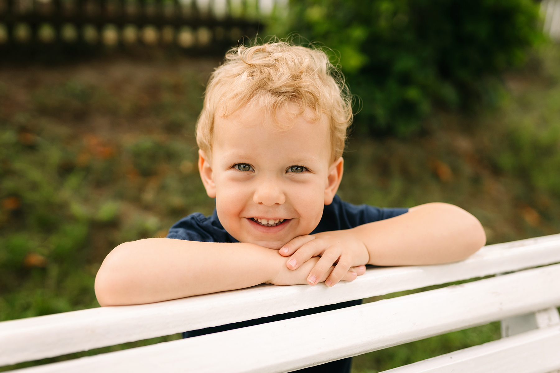 Blonder Junge mit lockigem Haar lehnt lächelnd auf einem weißen Gartenzaun