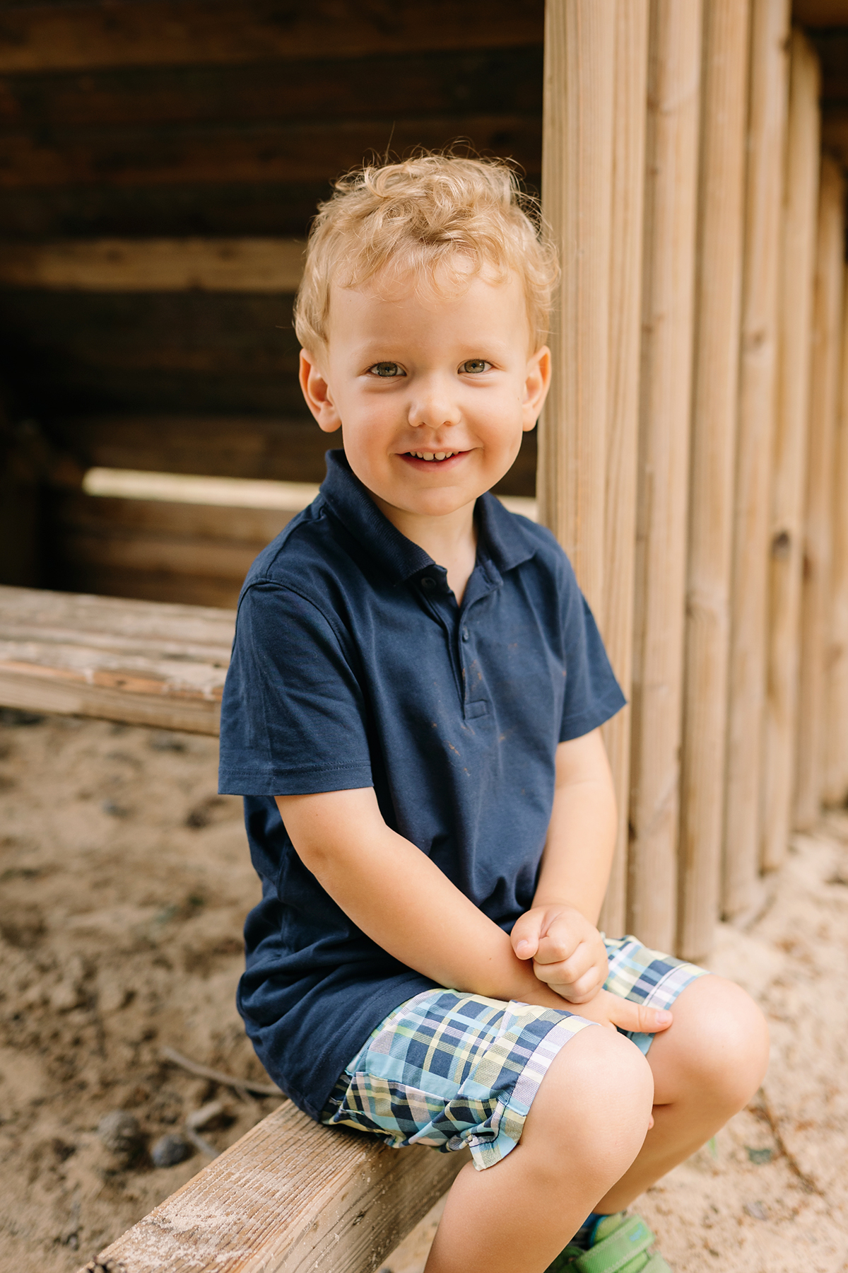 Blonder Junge in karierten Shorts sitzt lächelnd auf einem Holzbalken im Spielhaus